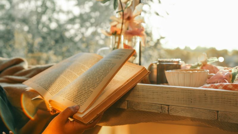 Person reading a book by a cozy window at sunset, with magnolia flowers, a black candle, and a cup of tea on a wooden tray. Warm golden light creates a peaceful, serene mood.