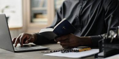 Person reading a Bible while working on a laptop, with a rosary and notebook nearby.