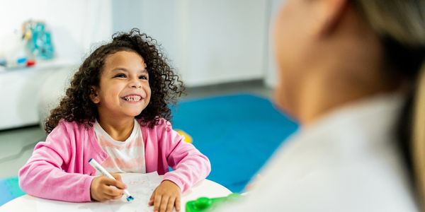 A smiling girl in a pink cardigan drawing with a marker at a table.