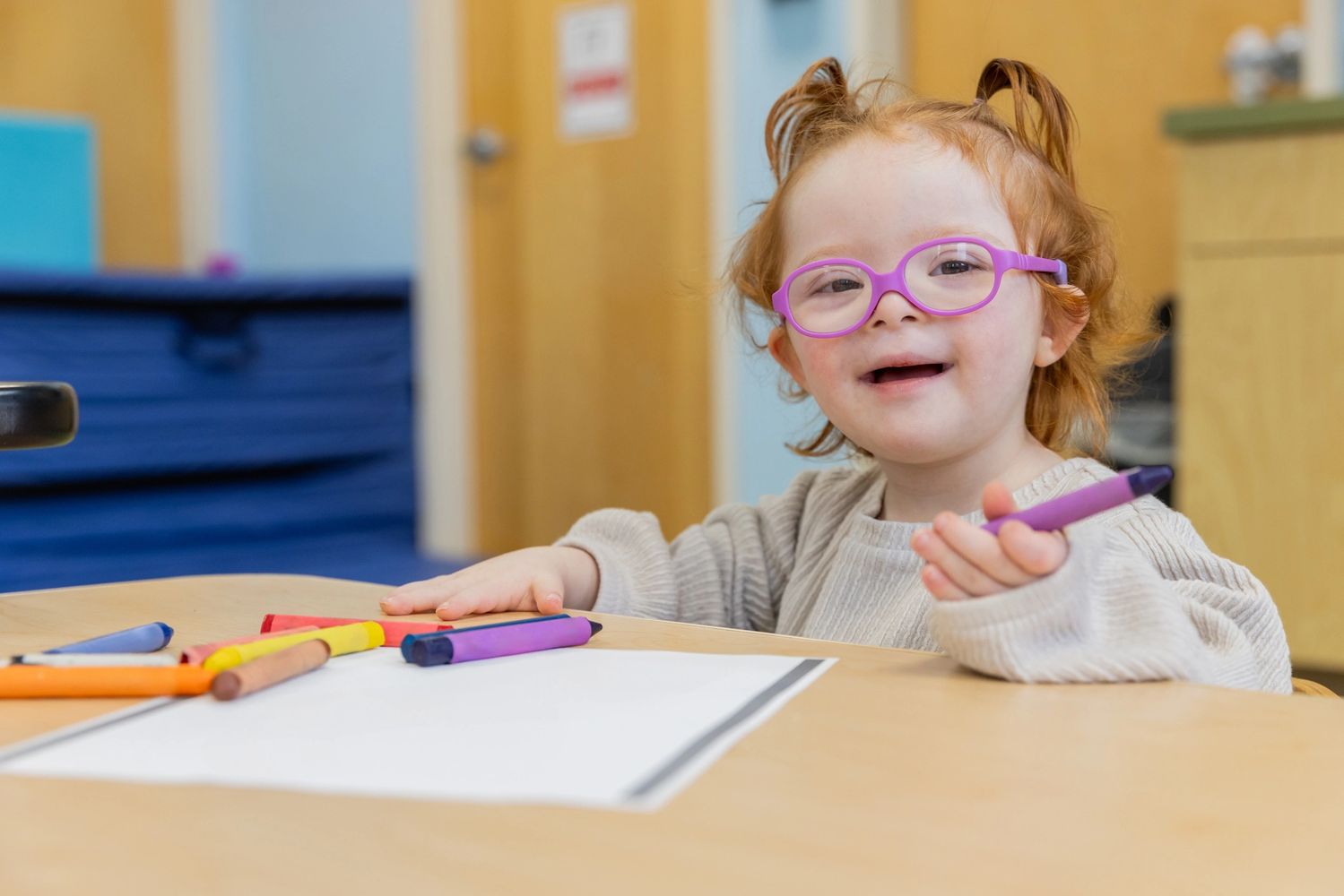 Smiling toddler with glasses holding a purple crayon at a table with crayons and paper.