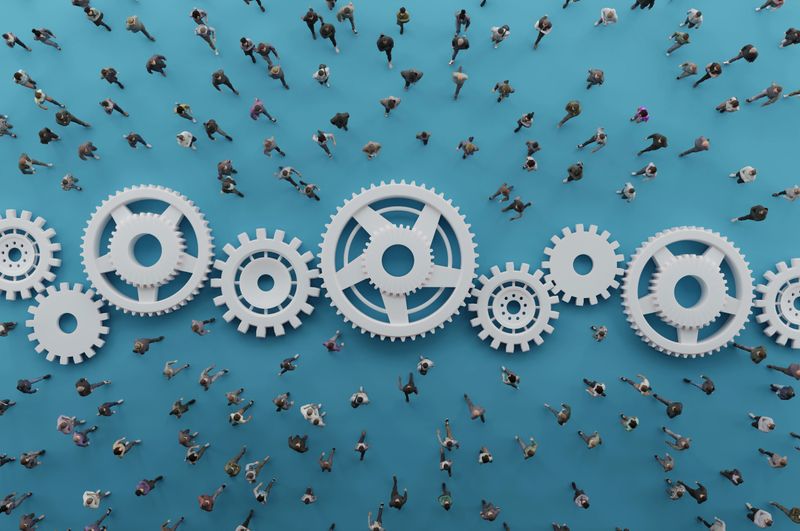 Overhead view of a diverse crowd walking across a bright blue surface interlinked with large white gears. The mechanical cogs symbolize teamwork, human systems, and the power of collective collaboration. Ideal for business, HR, and innovation themes.