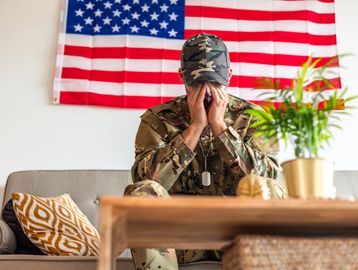 A soldier in uniform sits stressed with head in hands, an American flag behind.