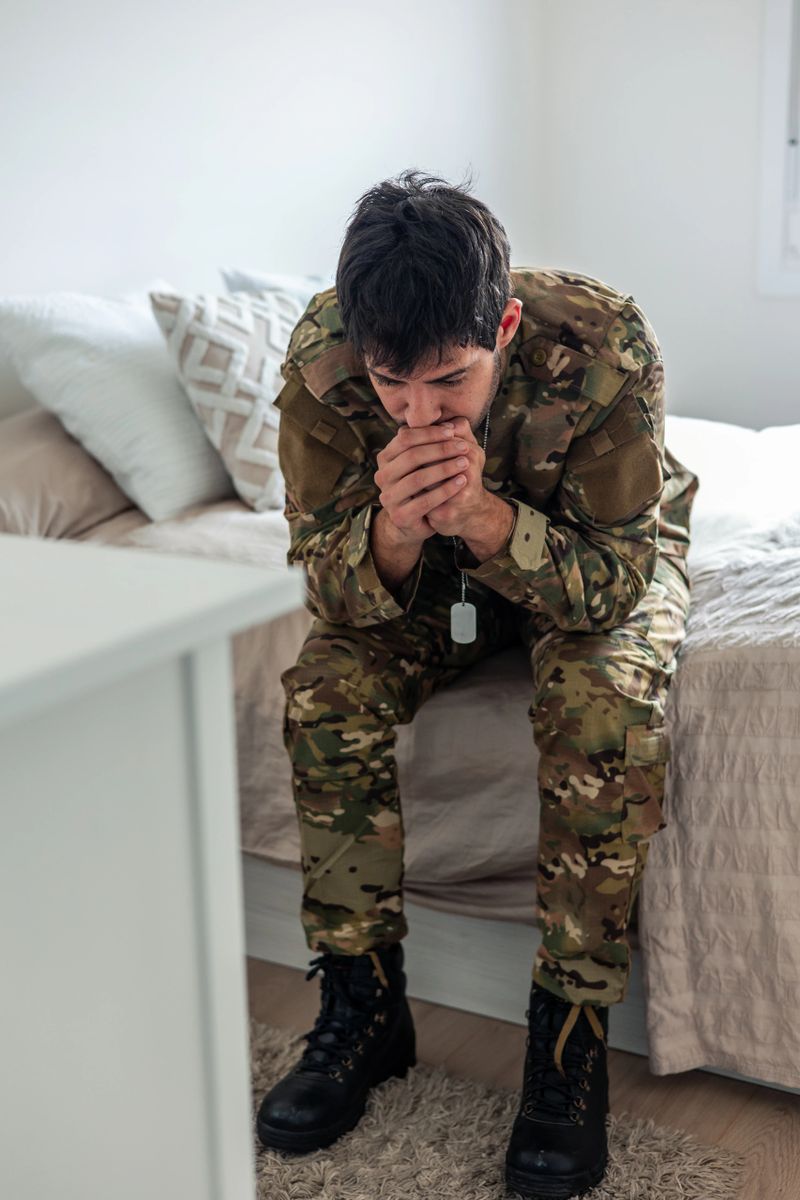 Young soldier in camouflage uniform sitting on his bed at home, hands clasped, suffering from post-traumatic stress disorder after returning from war