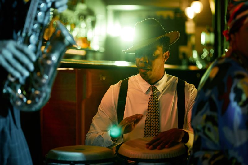 Medium shot of Black male drummer dressed in fashionable outfit with hat playing congas performing Afro Cuban jazz along with band in bar, under vibrant neon lights