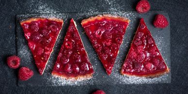 Four raspberry tart slices on a slate board with fresh raspberries.