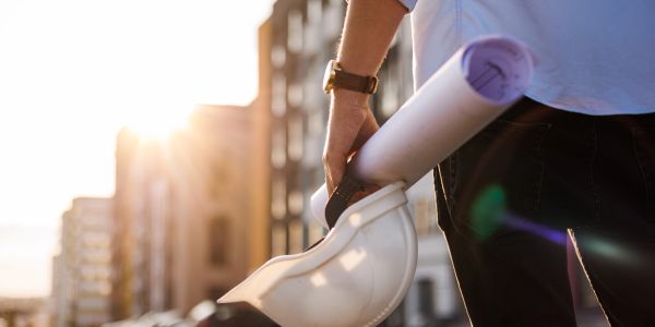 A person holding a white construction helmet and rolled blueprints at a building site during sunset.