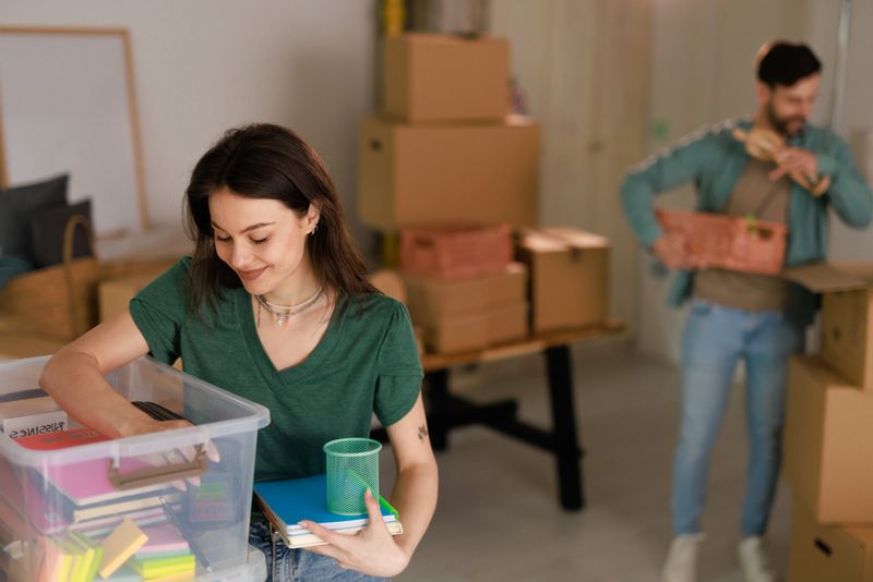 Young woman smiling while unpacking books and stationery from a plastic box, with her boyfriend carrying another box in the background, creating a cheerful scene of moving into a new home
