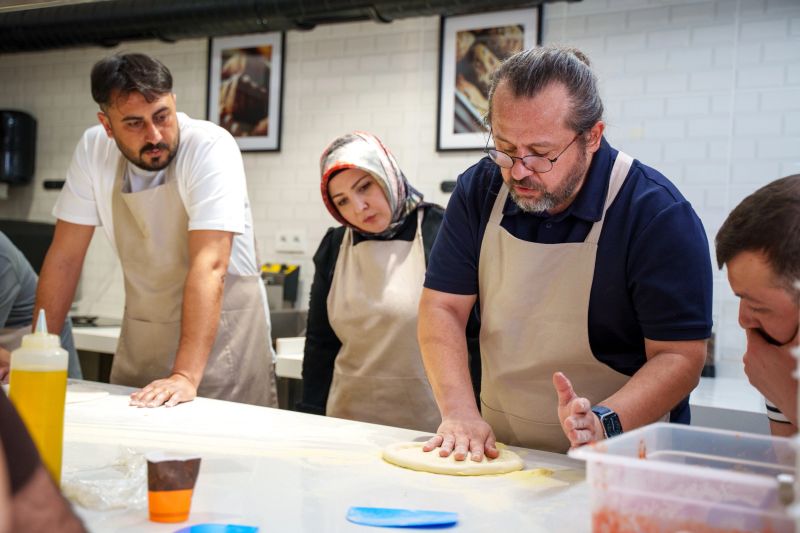 A diverse group of individuals wearing aprons are gathered around a white countertop, attentively watching a man demonstrate how to shape pizza dough during a cooking workshop. One woman in the group is wearing a hijab