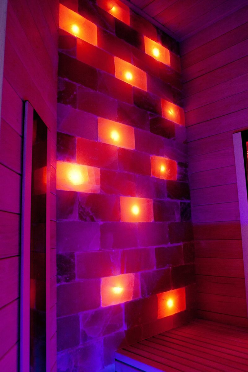 Infrared sauna room with illuminated Himalayan salt bricks emits a warm red and orange glow. Wooden bench and walls complement the relaxing ambiance. The salt bricks, set in a staggered pattern, create a calming visual effect, enhancing the therapeutic environment. Typically associated with wellness and detoxification, infrared saunas use heat to promote relaxation and health benefits. The warm lighting and natural materials contribute to a serene spa atmosphere, ideal for stress relief and rejuvenation.