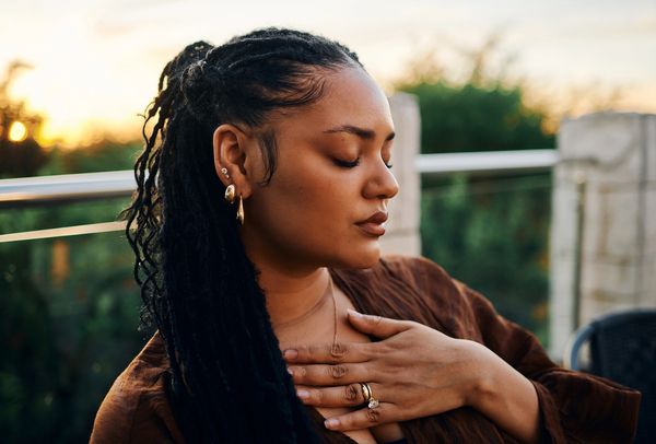 Woman with eyes closed, hand on chest, in peaceful outdoor setting at sunset.
