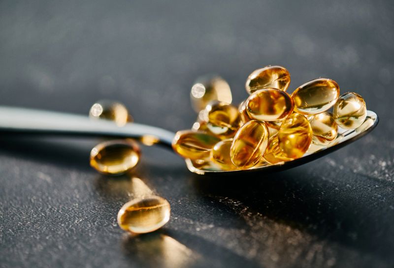 Close-up of shiny golden softgel capsules resting on a silver spoon, illuminated by soft light on a textured, dark surface.