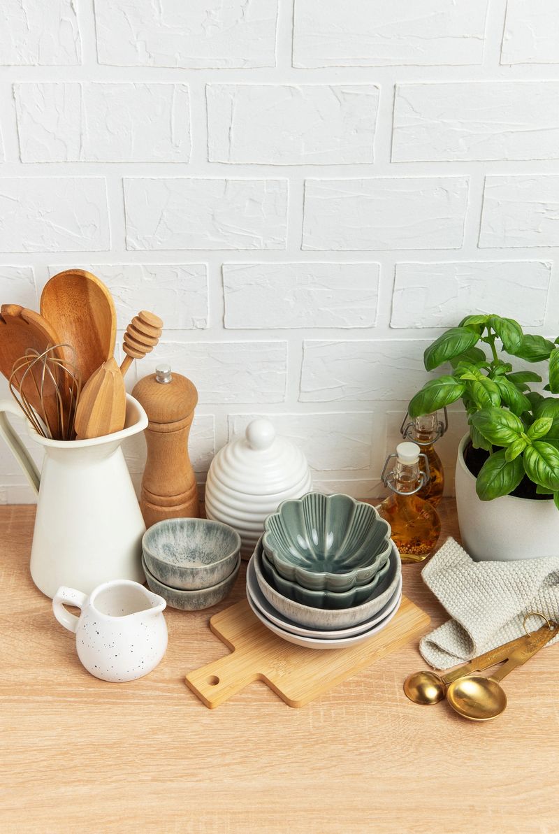 Various kitchenware, including bowls, utensils, spices, and a basil plant, arranged on a wooden countertop against a white brick wall