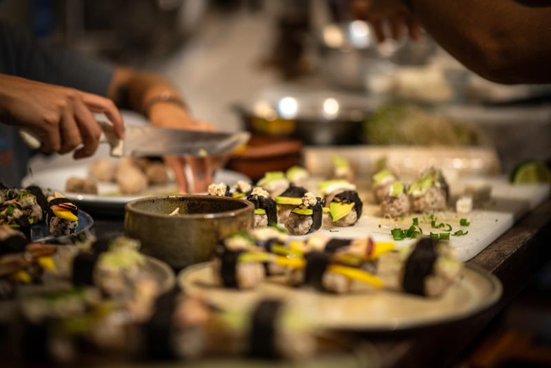 A close-up scene in a busy kitchen shows chefs preparing sushi rolls by hand. The foreground is filled with plates of colorful sushi garnished with avocado, mango, and seaweed. In the background, hands are actively slicing ingredients and assembling rolls.