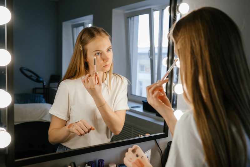 Young woman applying makeup with a focused expression in front of a mirror with vanity lights. She appears concentrated and engaged in refining her look in a well-lit room.