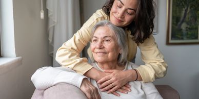 A young woman affectionately embraces an elderly woman on a couch indoors.