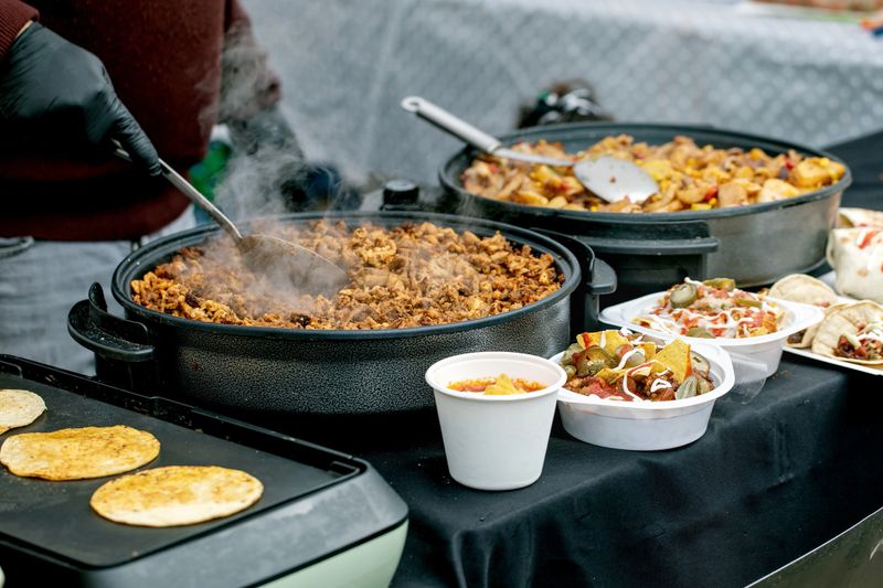Mexican street food preparation with cook stirring meat in cast iron pan. Tortillas taco ingredients sauces in containers ready for serving at outdoor street market.