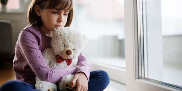 Sad girl hugging a teddy bear while sitting by a window.