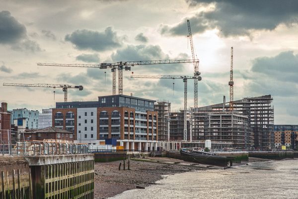 Urban waterfront under construction with cranes and buildings by the river.