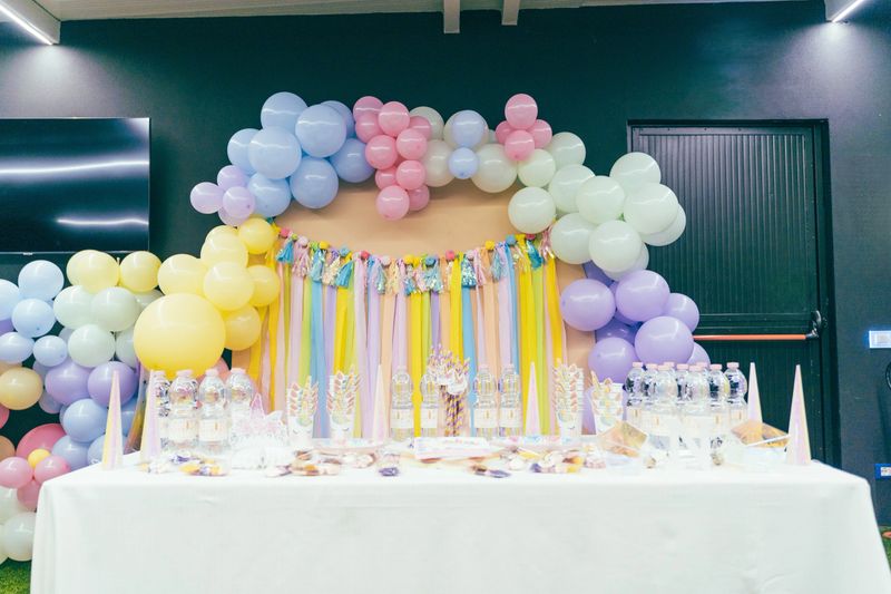Celebratory setup featuring a colorful balloon arch and birthday party table. Arrangement includes pastel balloons, tassel garland, bottled water, and party hats on a white tablecloth