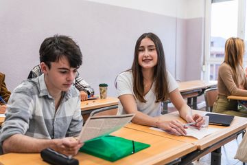Two students sitting at desks in a classroom, one reading and the other smiling.