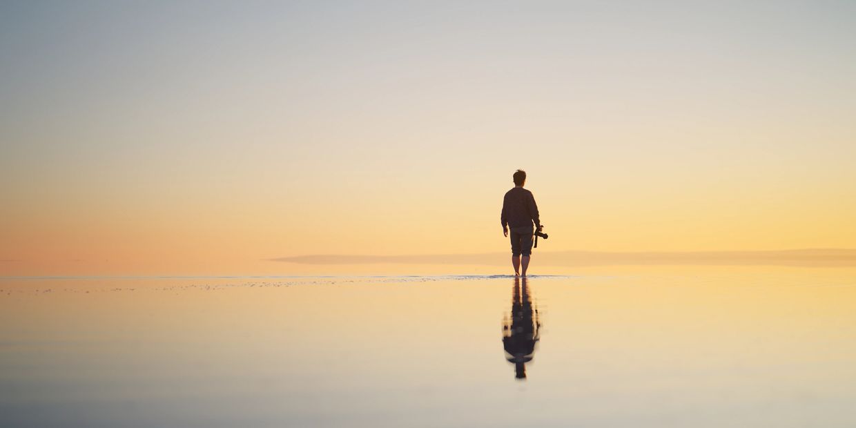 A person walks barefoot on shallow water at sunset holding a camera.