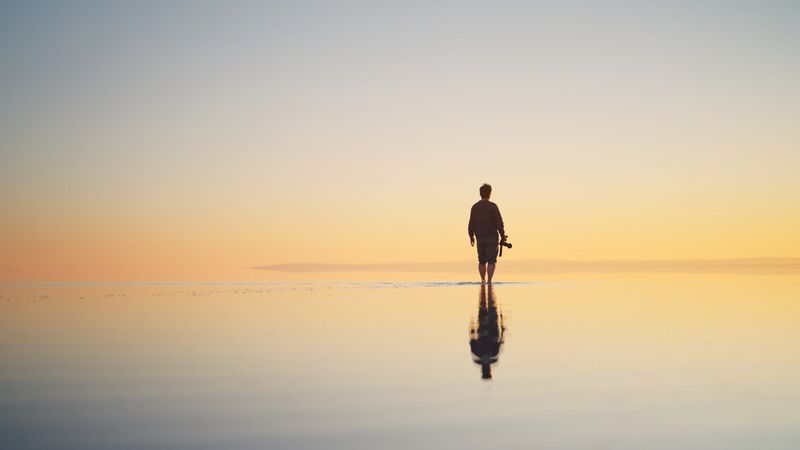 A rear view of a man solo traveler holding his camera and walking on the shallow surface of Salt Lake, Tuz Golu during sunset in Turkiye.