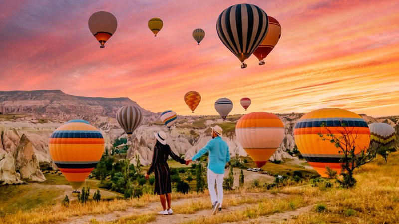 Mid-aged couple admirably gazes at the vibrant sunrise over Cappadocia. Hot air balloons fill the sky, creating a picturesque, romantic atmosphere in the unique landscape of Goreme.