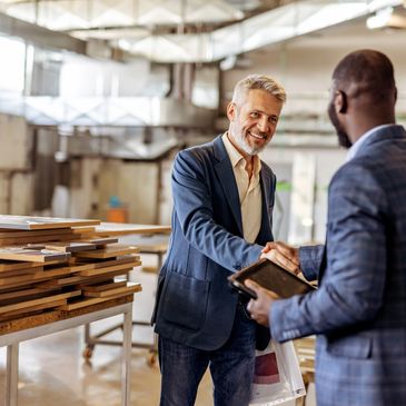 Two businessmen shaking hands in a woodworking shop with stacked wood panels.