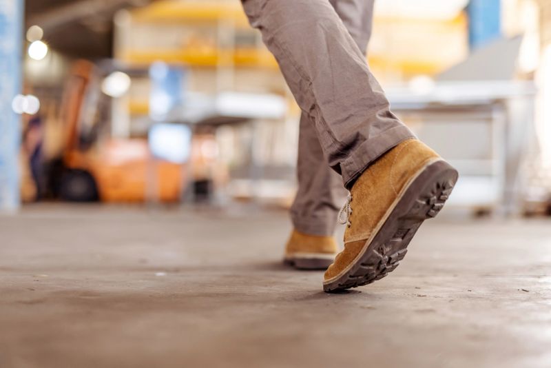 Worn leather boots stepping on a floor within a vibrant industrial setting, containing equipment and blurred background items. Captures movement and productivity in an active work location.