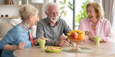 Drie oudere vrienden lachen rond de tafel met fruit en koffie.
