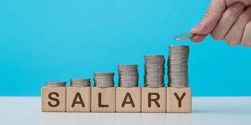 Stacked coins on wooden blocks spelling SALARY with a hand adding a coin.
