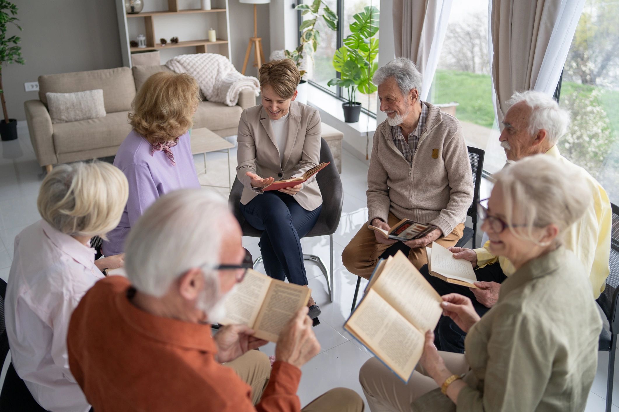 A group of seniors and a younger woman reading books together in a bright room.