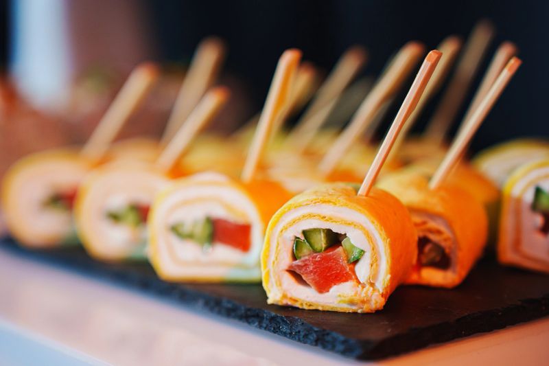 Various sushi rolls are arranged neatly on a black slate platter, showcasing vibrant colors and fresh ingredients. Chopped vegetables and seafood fill each delicate roll, appealing to the eye.