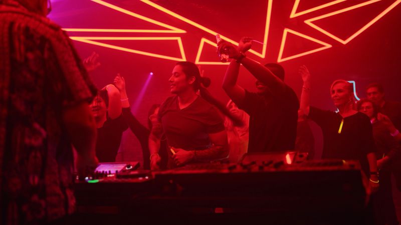 A DJ is playing music and a crowd of diverse people dancing in a nightclub. The crowd is diverse and there are people from different countries and different  ages.