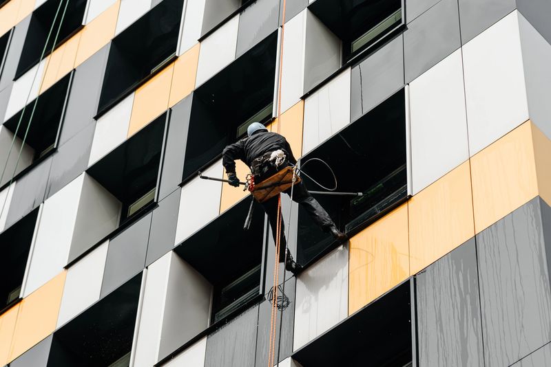 industrial climbers, window cleaner in helmet works on office building at height of several floors on cables, washing windows of metropolis business center with mops and pressure water machines