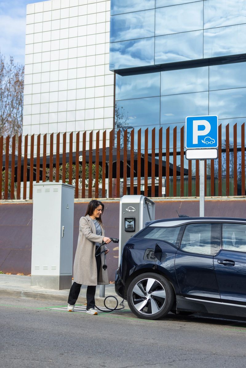 A woman charges her electric car at a public EV parking station, promoting sustainable mobility. Modern office buildings in the background. Vertical wide-angle shot with copy space.