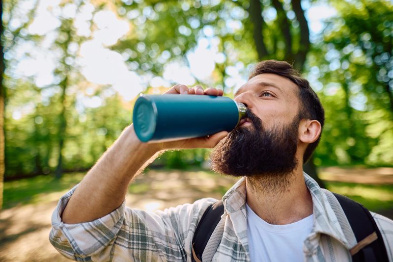 Bearded hiker enjoying a refreshing drink from a reusable water bottle while taking a break in a sunlit forest, surrounded by tall trees and the beauty of nature