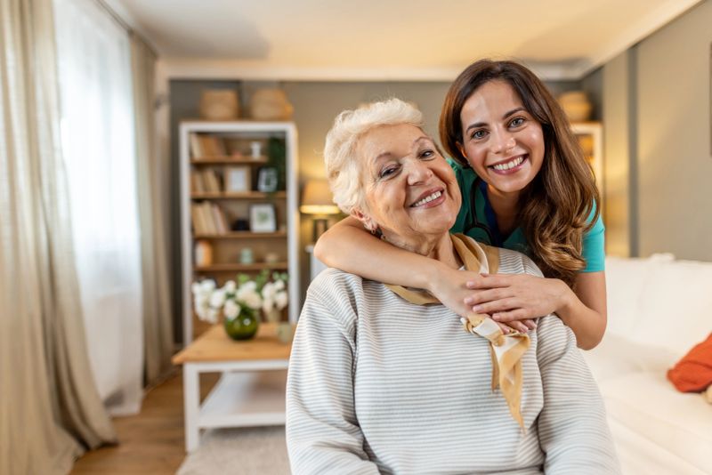A senior woman and her caregiver embracing warmly and smiling vividly in a well-lit living room, symbolizing care, companionship, and support. The scene captures the warmth of human connection and mutual respect.