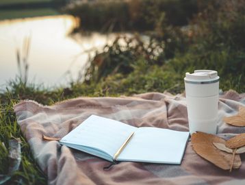 Notebook, pen, and travel mug on a blanket by the water.