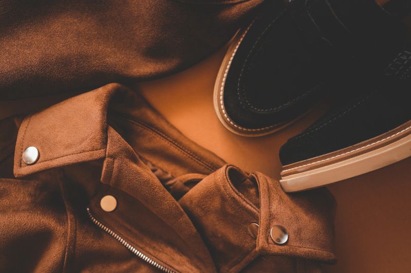 Flat lay of stylish brown suede jacket with metallic buttons and black suede shoes on a warm brown background, showcasing an elegant and modern autumn fashion outfit.