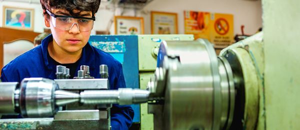 Young man operating a metal lathe in a workshop wearing safety goggles.