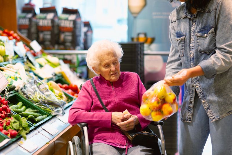 A young hispanic woman is helping a senior woman 93 years old, who is sitting in a wheelchair, with her purchase, showing a plastic bag with apples