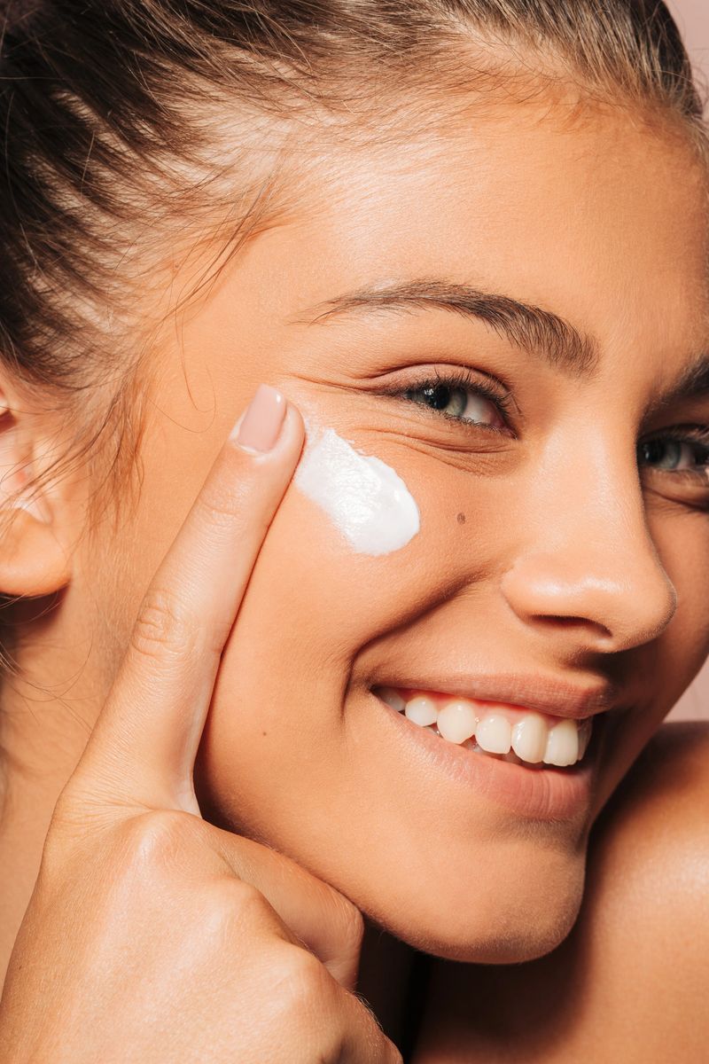 This is a close-up photo of a smiling young woman applying face cream to her cheek. She has clear, glowing skin and is gently rubbing the cream in with her finger. Her hair is tied back, and the background is a soft pink color. The image looks fresh and natural, showing a skincare routine. Studio shot
