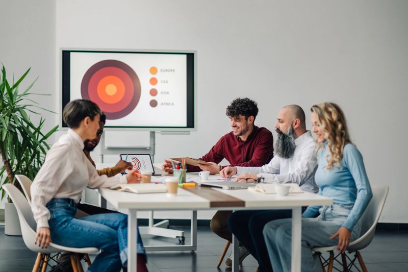Five business professionals are having a productive meeting, analyzing global market data displayed on a presentation screen