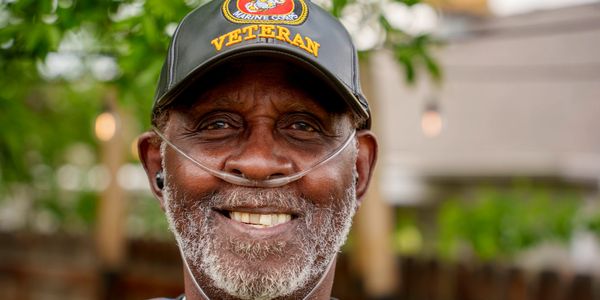 Smiling elderly man wearing a Marine Corps veteran cap with oxygen tube.