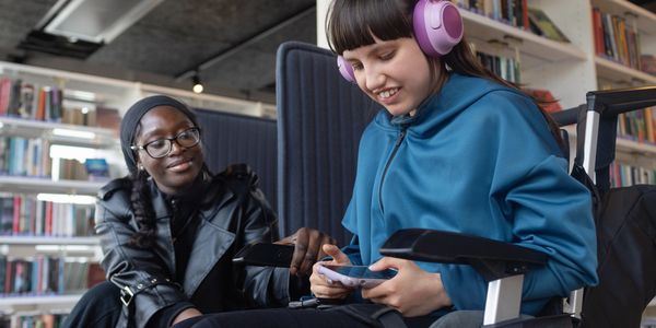 Two girls sharing a moment in a library, one in a wheelchair.