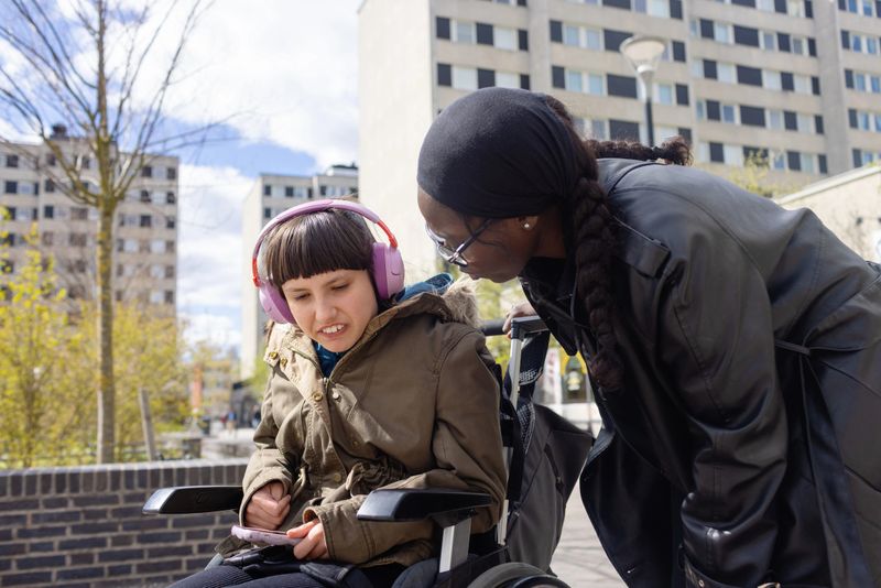 A neurodivergent teenage girl with autism and Rett syndrome sits in a wheelchair wearing pink headphones in a sunny outdoor setting. Her disability support worker leans over her to talk.