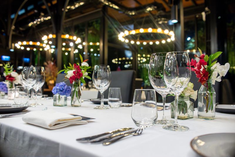 A luxurious dinner table setup in a modern venue with grand lighting and floor-to-ceiling windows. Floral centerpieces and polished glassware enhance the upscale, sophisticated atmosphere.