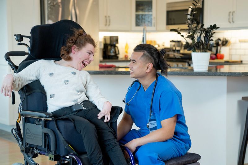 A caregiver in scrubs assists a smiling young woman in a wheelchair, communicating warmly. The setting includes a modern kitchen, reflecting care, support, and inclusivity in a home environment.