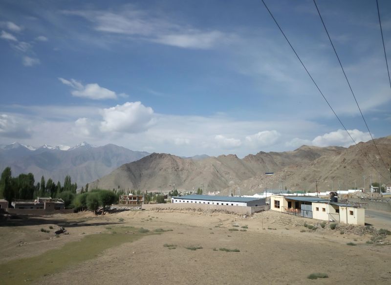 Vast barren land near Leh Ladakh features mountains, small villages, and lush trees with clouds scattered in the clear sky.